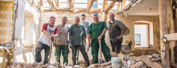 In einem sanierungsbedürftigen Haus mit offenem Dach und eingerissenen Wänden stehen sechs Männer, mit Werkzeugen in der Hand