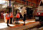 Auf diesem Foto sind 4 Personen zu sehen die in einer Werkstatt für Güterzüge stehen und sich unterhalten. Drei davon tragen orangene Warnwesten. 