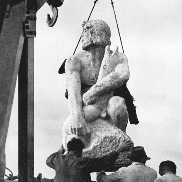 Eine große Statue aus Stein (Ein Flussgott, der einen großen Fisch im Arm hält) wird von einem Kran abgeladen. Drei Männer begleiten die Lieferung