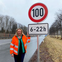 Verkehrsministerin Kerstin Schreyer steht mit oranger Warnweste neben einem Verkehrsschild an einer Autobahn. Das Verkehrsschild gibt ein Tempolimit von 100 km/h im Zeitraum von 6 bis 22 Uhr vor.  © StMB