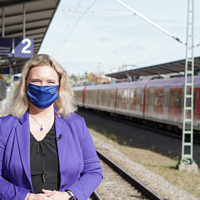 Verkehrsministerin Kerstin Schreyer steht mit Maske an einem Bahnsteig. Im Hintergrund ist ein Zug zu sehen.