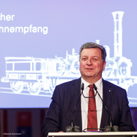 Verkehrsminister Christian Bernreiter steht an einem Rednerpult und hält eine Rede. Im Hintergrund eine Leinwand mit Aufdruck der Adler-Lok und Text „Bayerischer Eisenbahnempfang 2022“. © Daniel Karmann