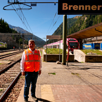 Hier steht Herr Staatsminister Christian Bernreiter an einem Gleis am Brenner. Der Himmel ist blau. Im Hintergrund sind Gleise und ein roter Zug zu sehen.  © StMB