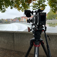 Eine Kamera auf einem Stativ steht an einem Flussufer. Im Hintergrund ist eine Stadt zu sehen.